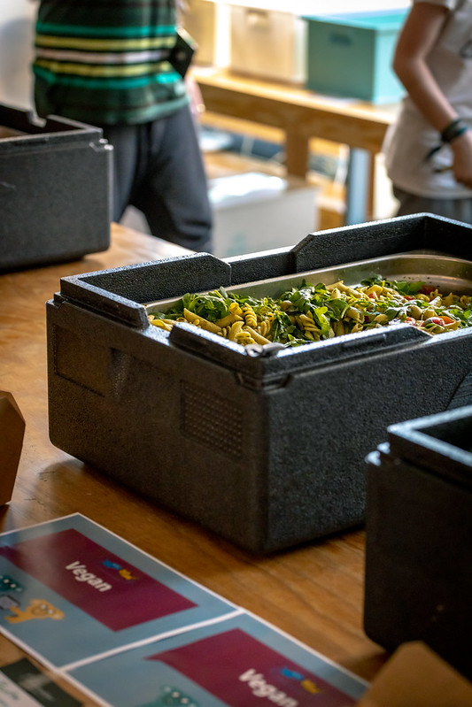 Symbolbild für gesunde Verpflegung.
Auf dem Tisch steht ein Gastrobehälter mit einem veganen Nudelgericht in einer Warmhalte-Box. Davor liegt ein Zettel mit der Aufschrift “vegan”