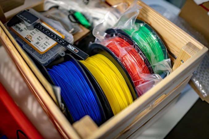 Symbolbild für Verbrauchsmaterial
Eine Holzkiste mit Rollen mit farbigem (blau, gelb, rot, grün) Filament für 3D-Drucker