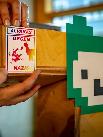 Symbolbild Dekoration. Auf ein Rednerpult mit Jugend hackt Logo wird ein Sticker mit dem Text “Alpakas gegen Nazis” in Regenbogenfarben geklebt