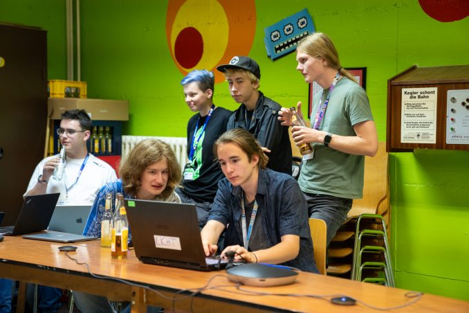 Eine Gruppe Jugendliche von 6 Personen bei der Projektarbeit. Zwei sitzen konzentriert vor einem Laptop. Die Anderen unterhalten sich im Hintergrund